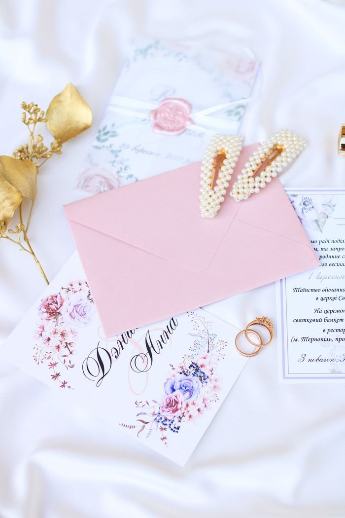 services-01 Flat lay of a wedding invitation with floral design, pink envelope, pearl hair clips, and rings.