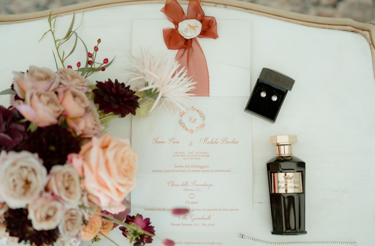 who-we-are A stylish flat lay featuring a wedding invitation, flowers, perfume, and jewelry with a classic touch.