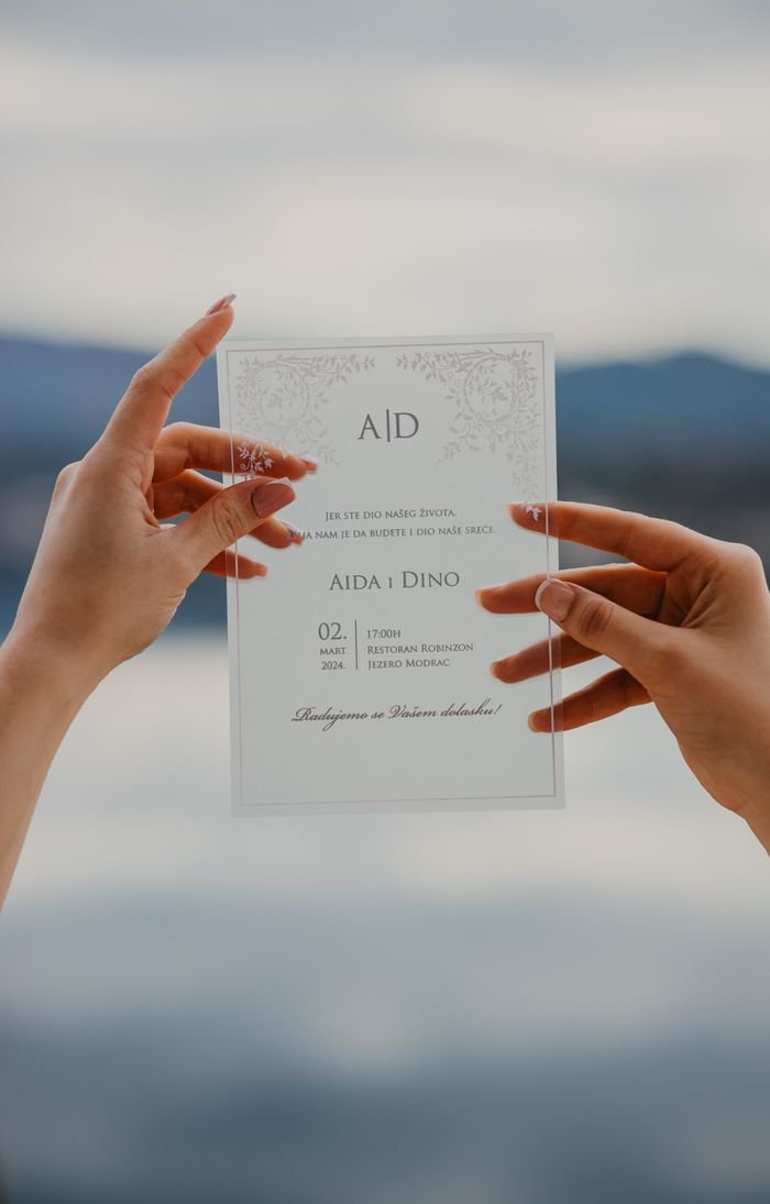 A transparent wedding invitation card held by two hands against a scenic background.